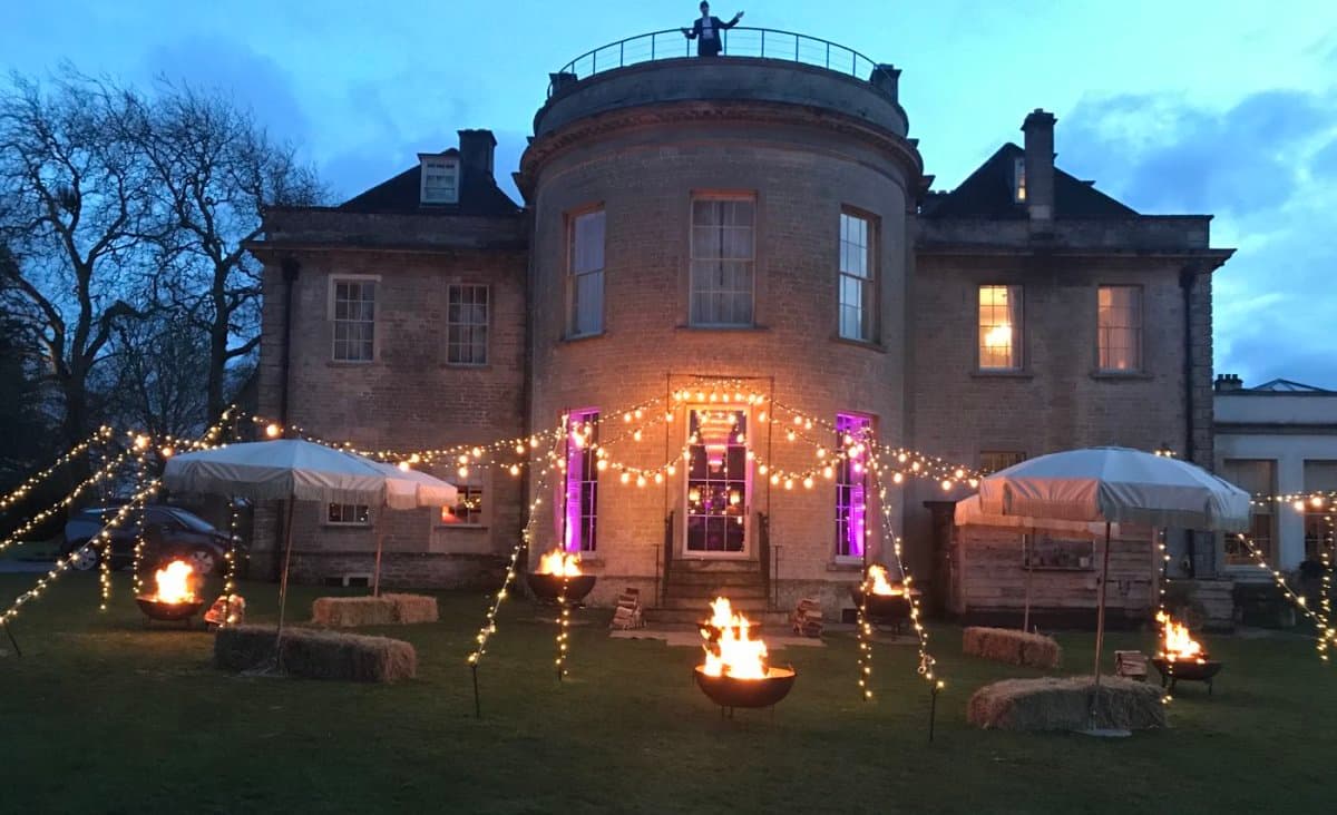 Festoon and fairy-light fan over a lawn with multiple fire pits at dusk