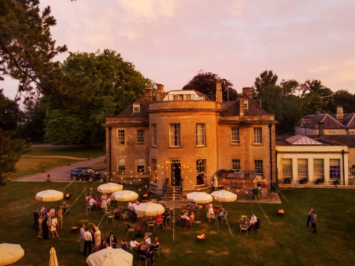 Manor house lawn at dusk with festoon lighting and multiple fire pits among guests