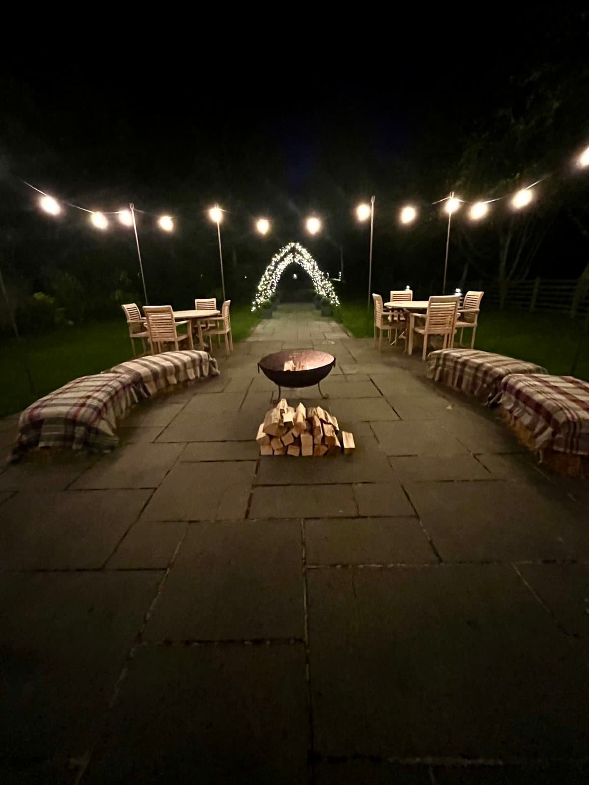 Fire pit on a stone patio with festoon lighting, hay-bale seating and plaid blankets