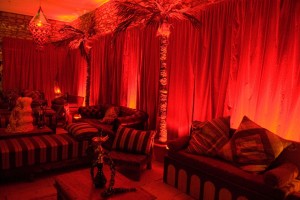 A Moroccan themed party space. Red uplighting, fake palm trees, low furniture and shisha pipes all displayed.