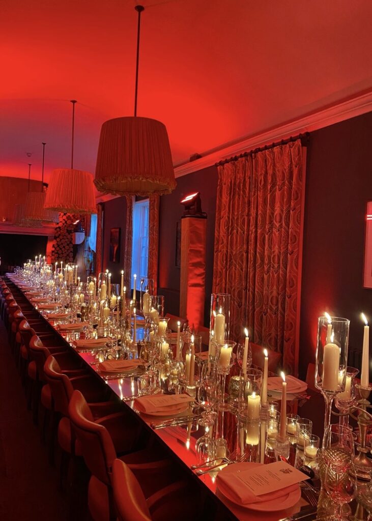 Red themed dinner party with a mirror table top with many candles on the table and red lighting throughout, with red chairs down one side of the table.