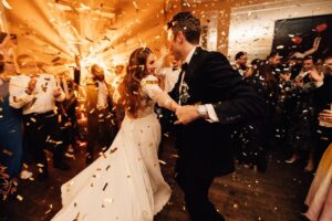 A bride and groom dancing their first dance surrounded by clapping people, confettit, in the air, dance-floor lighting behind the happy couple.