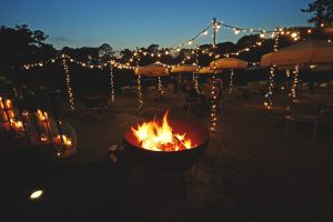 Fire Pit in foreground at dusk wit a lighting canopy of fairy-light behind the fire-pit with cream lawn umbrellas.