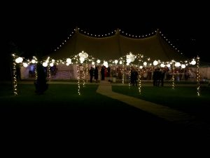 A canvas marquee at night set on a lawn with a free standing lighting canopy with fairy-lights and white shades