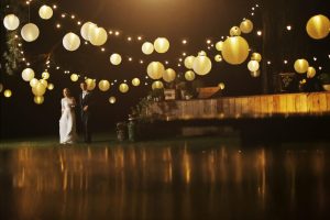 Nightime, exterior, many white shades hanging in the air from festoon lightng. Bride & Groom to the left smiling.