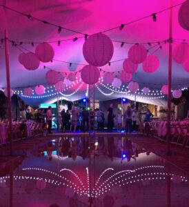 A stretch marquee with lighting design of pink mood lighting and festoon with the lights reflected in the water below and people in the distance dancing