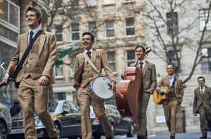 Five men in check suits walkign down a London road with instruments in hand