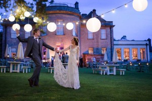 A bride and groom hand in hand outside at dusk. They are underneath a tree with festoon lighting and white shades above. Behind i an english, country house hotel