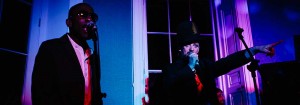 Boy George singing into a microphone whilst pointing and a man to the left singing with pink mood lighting a window frame.