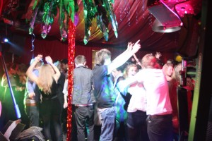 Young people dancing at a party with arms raised. A red fabric ceiling is above and fake, plastic palm trees in the foreground