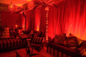A Moroccan Themed room that has been re-styled with red lighting, morrocan low furniture, shisha pipes on loww wooden tables and 2 fake Palm trees.