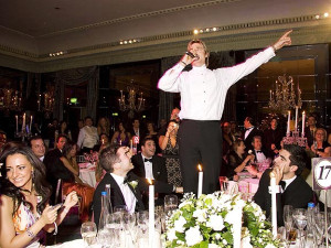 The singing waiters performing in a large dining room with the perfomer stading on the table and everyone smiling and looking at him.