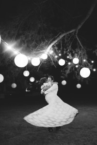 A bride and groom in a passionate embrace under a tree with festoon lighting and white shades in this black and white image