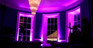 3 windows with pink lighting their white frames with a chandlaier visible which is triangle shaped