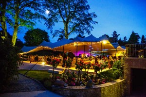 Outdoor at night with a stretch marquee and 2 large trees in the background. Inside the marquee people are gathered around tables.