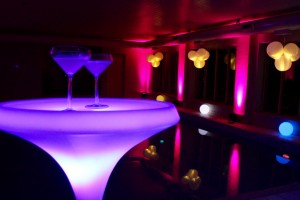 A swimming pool at night with pink lighting up coloums and a led cocktail table in the foreground with 2 cocktail glasses.