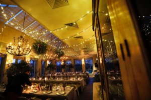 A large dining room with 3 tables set for a wedding with a fiary-light canopy overhead and chandaliers glowing