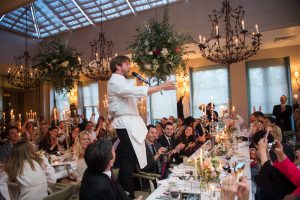 A singing waiter standing on a chair with lots of smiling people looking at him whilst he is singing at a wedding breakfast