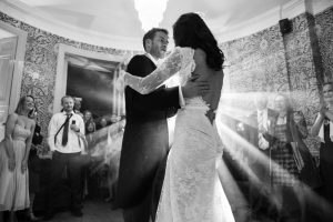A bride and groom perofrming their first dance with white lighting behind them and smiling people around the edges of the rooom