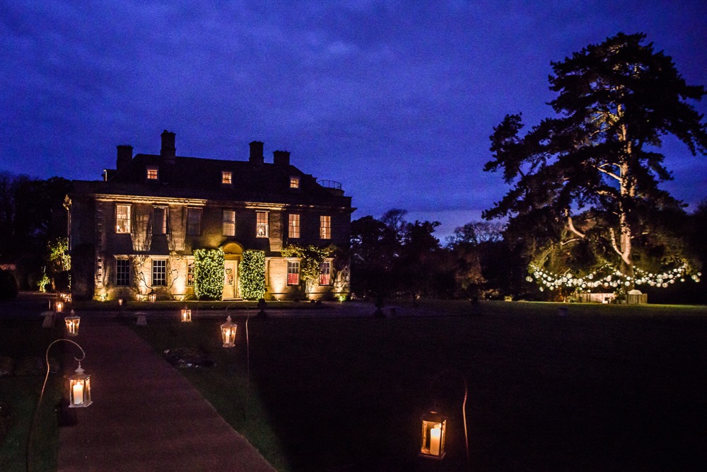 Dusk, hotel in background, large tree to the right with festoon lighting and white shades, lanterns and crooks in foreground leading to the hotel.