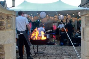 A large fire-pit burning with a chef with tongs behind a jazz band playing framed by 2 stone pillars.