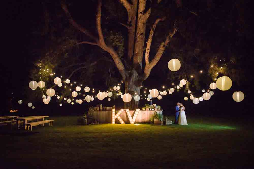 A bride and groom under a large pine tree with a K & V light up sign and a stunning lighting canopy lighting thme and the lawn