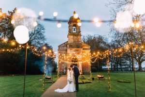 Church in background with a fairy-light tunnel in the foreground with a bride and groom embracing in the centre