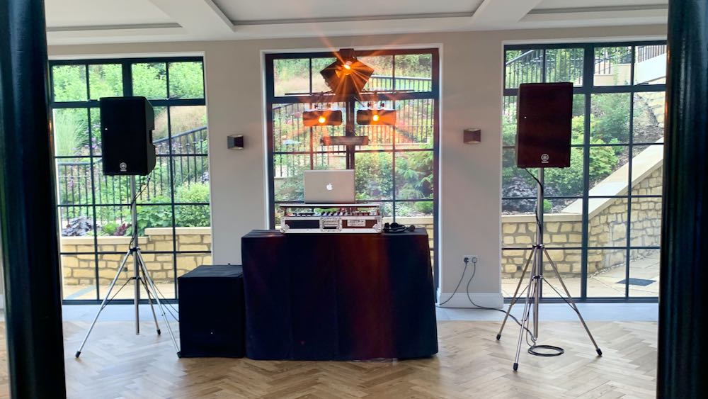 DJ Setup, daytime, large windows behind DJ equipment, pair of speakers left and right of sj decks, lighting stand behind in centre.