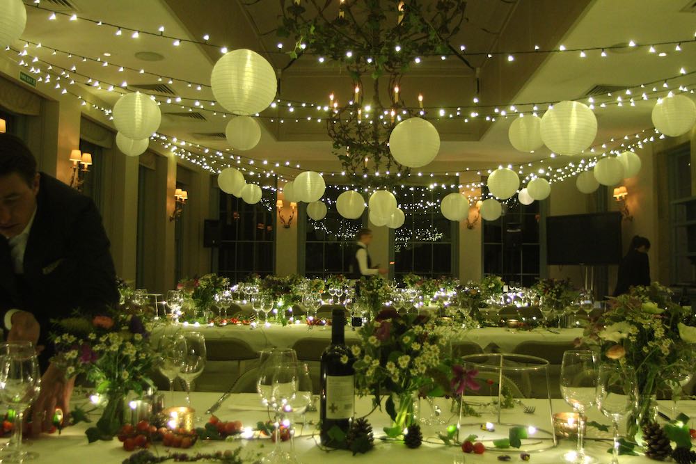 Dining room, night, tables left to right, flowers in vases, fairy-light canopy with white shades attached. Windows in distance reflecting the fairy-lights.