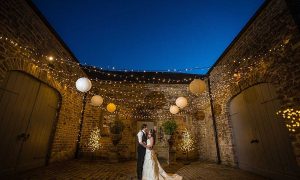 A bride and groom embrace under a fairy-light canopy with white shades. Surrounded bya 3 sided stone building at dusk.