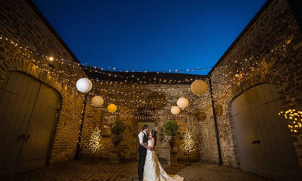A bride and groom embrace under a fairy-light canopy with white shades. Surrounded bya 3 sided stone building at dusk.