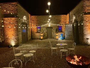 Night, 2 sided stone building, amber lighting in corners, white tables & chairs in foreground on gravel , fire-pit lit in right front.