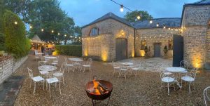 Dusk, exterior, background 3 sides stone building, foreground, fire-pit, gravel, white table and chairs, above a festoon lighting canopy
