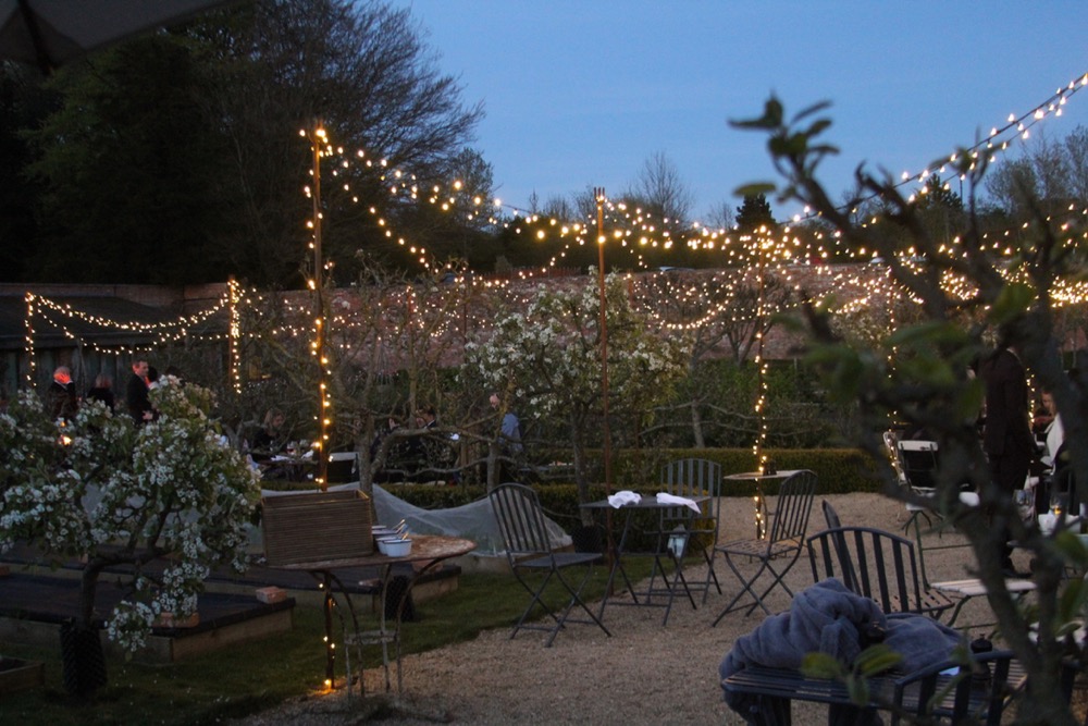 Walled Garden at dusk, fairy-lights run through trees and bushes. gravel on floor with tables and chairs