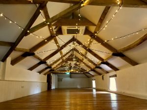 An empty barn room with wooden floor, 2 small windows on the rght, fairy-light canopy suspended from beams