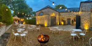 Stone coach house in background, gravel in foreground with fire-pit, white metal tables and chairs. A festoon ligting canopy stretches to the pizza tent in the far distance.