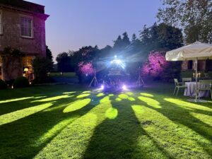 DJ Equipment setup on a croquet lawn on a sunny night. House to the left and an umbrella and table to the right.