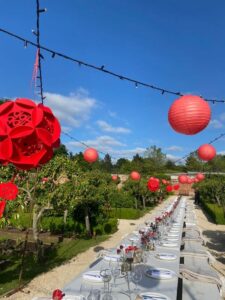 A long table in a walled garden with beautiful blue sky and a number of red shades suspended from a fairy-light canopy