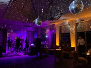 2 SIlver Mirrorballs suspended from the ceiling with 2 chandaliers either side. A band is preparing below at one end and the room is light with amber and pink lighitng.