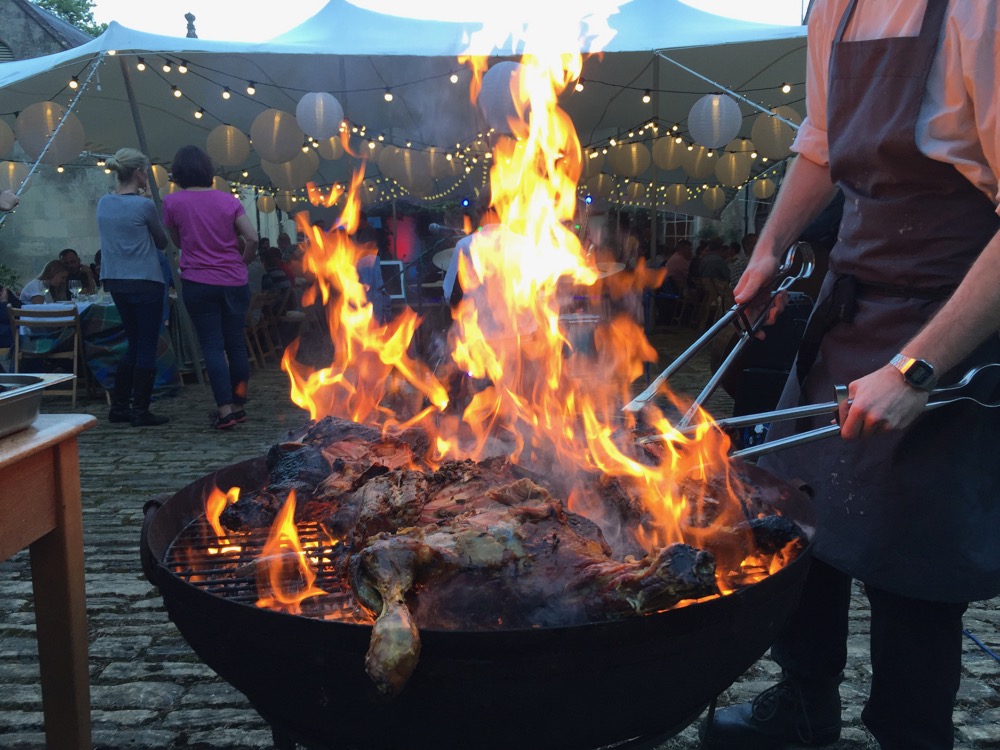 Meat being cooked on a large fire-pit with huge flames and a stretch marquee in the background.