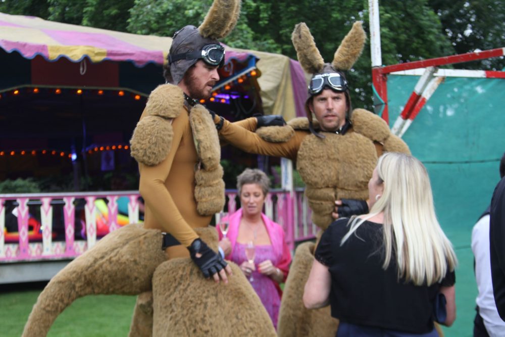 A lady talking to giant human Kangaroos with Dodgems in the background. The kangaroos include leather flying helmets.